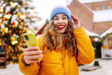 Arka planda dekoratif Noel ışıkları olan akıllı telefonu kullanan genç bir kadın. Noel Fuarı.