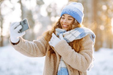 Gülümseyen kadın selfie çekiyor ve kış parkında el sallıyor. Blog yazma, seyahat, hafta sonu kavramı. Etkin yaşam biçimi.