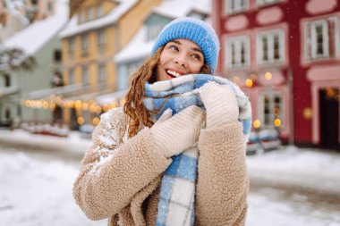 Gülümseyen kadın kışın sokakta kareli atkı, ceket ve örgü şapka giyerek yürüyor. Mutlu ruh hali. Moda tarzı trend. Kış tatili.