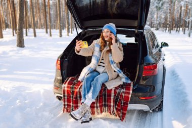 Young Woman sitting in the trunk of her car and taking selfie on a background of snow-covered winter forest. Concept of technology, adventure, travel by car.