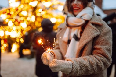 Noel parıltılarını ellerinle kapat. Soğuk kış akşamında Noel pazarında elinde yanan maytap tutan genç bir kadın. Noel, yeni yıl.