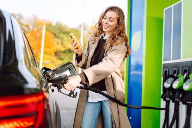 Benzin istasyonunda arabasını benzinle dolduran güzel bir kadın. Çevrimiçi ödeme. Kadın, telefonundaki uygulamayı kullanarak benzin istasyonunun parasını ödüyor..