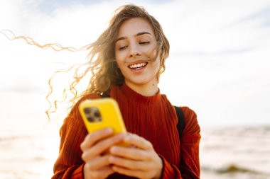 Plajda cep telefonu kullanan genç bir kadın. Soğuk hava. Sonbahar. Bloglama, iletişim, teknoloji kavramı.