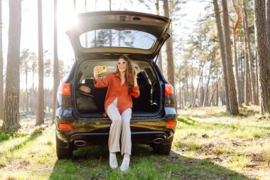 Neşeli bir kadın arabasının bagajında oturup selfie çekiyor. Arabayla seyahat ederken selfie. Seyahat, hafta sonu.