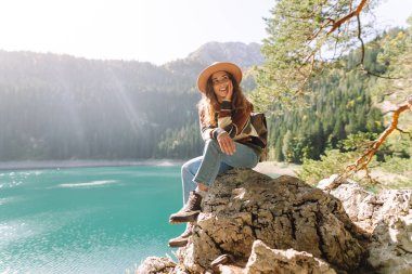 Yalnızlık zamanı. Mutlu turist kadın güneşli havada dağ gölü manzarasının tadını çıkarıyor. Dağlarda bir kayanın üzerinde oturan bir kadın, bir yürüyüşün ortasında dinleniyor..
