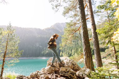 Yalnızlık zamanı. Mutlu turist kadın güneşli havada dağ gölü manzarasının tadını çıkarıyor. Dağlarda bir kayanın üzerinde oturan bir kadın, bir yürüyüşün ortasında dinleniyor..