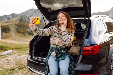 Araba yolculuğu ile selfie. Kafkasyalı genç bir kadın doğada bir arabanın bagajında otururken akıllı telefon kullanıyor..