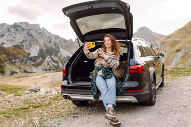 Araba yolculuğu ile selfie. Kafkasyalı genç bir kadın doğada bir arabanın bagajında otururken akıllı telefon kullanıyor..