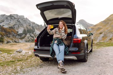 Araba yolculuğu ile selfie. Kafkasyalı genç bir kadın doğada bir arabanın bagajında otururken akıllı telefon kullanıyor..
