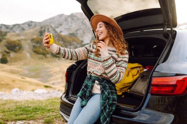 Araba yolculuğu ile selfie. Kafkasyalı genç bir kadın doğada bir arabanın bagajında otururken akıllı telefon kullanıyor..