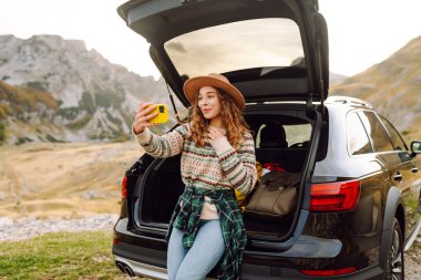 Araba yolculuğu ile selfie. Kafkasyalı genç bir kadın doğada bir arabanın bagajında otururken akıllı telefon kullanıyor..