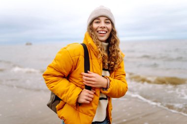 Sarı ceketli güzel bir kadın rüzgarlı bir sonbahar gününde sahilde dinleniyor. Yaşam tarzı, seyahat, turizm, doğa, aktif yaşam.