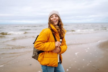 Sarı ceketli güzel bir kadın rüzgarlı bir sonbahar gününde sahilde dinleniyor. Yaşam tarzı, seyahat, turizm, doğa, aktif yaşam.