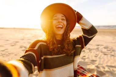 Genç bir kadın gün batımında kumlu bir sahilde neşeli bir selfie çekiyor. Blogculuk, teknoloji, yaşam tarzı, seyahat, turizm, aktif yaşam.