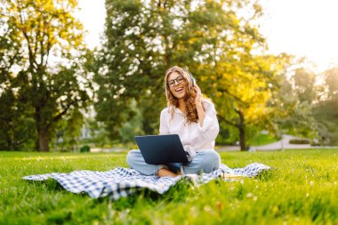 Dizüstü bilgisayarı ve kulaklığı olan genç bir kadın parkta yeşil çimlere oturmuş çalışıyor. Müzik konsepti. Eğitim, iş, blog veya serbest çalışma kavramı.