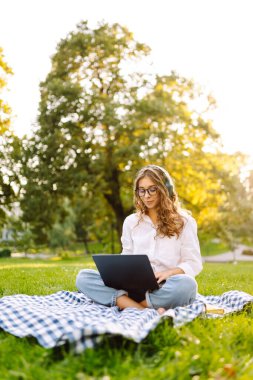 Dizüstü bilgisayarı ve kulaklığı olan genç bir kadın parkta yeşil çimlere oturmuş çalışıyor. Müzik konsepti. Eğitim, iş, blog veya serbest çalışma kavramı.