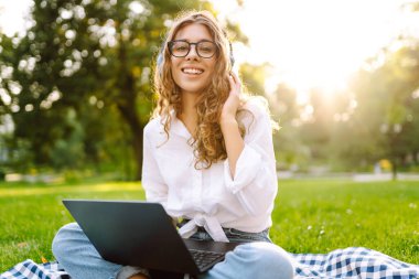 Dizüstü bilgisayarı ve kulaklığı olan genç bir kadın parkta yeşil çimlere oturmuş çalışıyor. Müzik konsepti. Eğitim, iş, blog veya serbest çalışma kavramı.