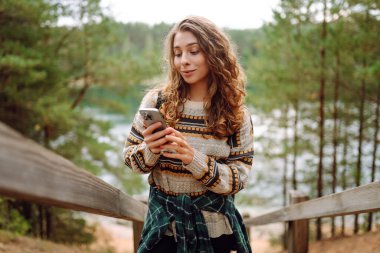 Güzel bir göl kenarında, rahat bir kazak giymiş gülümseyen ve telefonunu kullanan genç bir kadın. Macera, doğa, blog yazma.