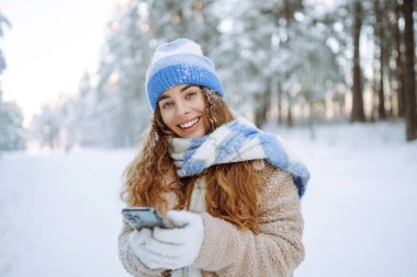 Sıcak kıyafetler içinde mutlu bir kadın kış ormanlarında kar kaplı ağaçlara ve düşen kara karşı akıllı telefonuyla selfie çekiyor. Kış tatili.