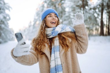 Sıcak kıyafetler içinde mutlu bir kadın kış ormanlarında kar kaplı ağaçlara ve düşen kara karşı akıllı telefonuyla selfie çekiyor. Kış tatili.