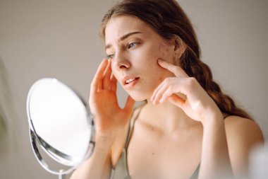 Güzel bir kadın aynaya bakıyor, eliyle yüzüne dokunuyor. Sivilceler, sivilceler, gözenekler, yaralar. Filtresiz doğal cilt