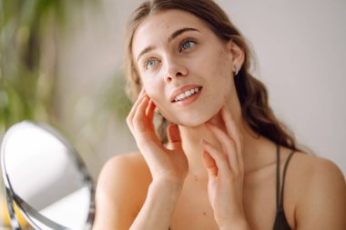 Güzel bir kadın aynaya bakıyor, eliyle yüzüne dokunuyor. Sivilceler, sivilceler, gözenekler, yaralar. Filtresiz doğal cilt