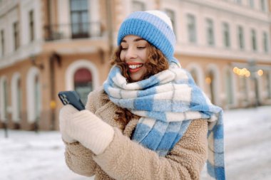 Caddede yürüyen çekici bir kadın ve karlı şehir merkezinde akıllı telefondan mesaj atıyor. Çevrimiçi iletişim. Teknoloji konsepti.