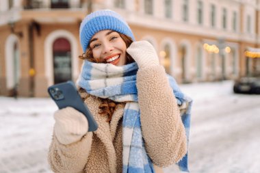 Caddede yürüyen çekici bir kadın ve karlı şehir merkezinde akıllı telefondan mesaj atıyor. Çevrimiçi iletişim. Teknoloji konsepti.