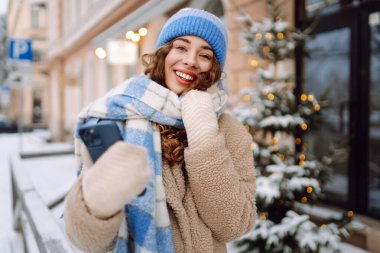 Caddede yürüyen çekici bir kadın ve karlı şehir merkezinde akıllı telefondan mesaj atıyor. Çevrimiçi iletişim. Teknoloji konsepti.