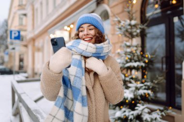 Caddede yürüyen çekici bir kadın ve karlı şehir merkezinde akıllı telefondan mesaj atıyor. Çevrimiçi iletişim. Teknoloji konsepti.