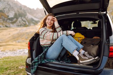 Genç bir kadın bir arabanın bagajında oturuyor. Sonbahar manzarası. Arabayla seyahat etme kavramı, aktif yaşam tarzı..