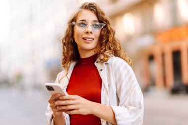 Şehirde sokakta yürürken telefonunu kullanırken gülümseyen çekici bir kadın. Teknoloji, blog yazma, iletişim kavramı.