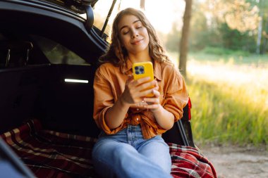 Mutlu kadın dışarıda bir arabanın bagajında oturuyor ve akıllı telefon kullanıyor. Arabayla seyahat etme kavramı, aktif yaşam tarzı..