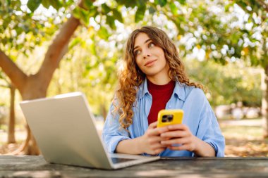 Genç gülümseyen öğrenci bahar parkında bankta oturur, dinlenir, dizüstü bilgisayar kullanır, cep telefonu interneti tarar. Kentsel yaşam tarzı kavramı.