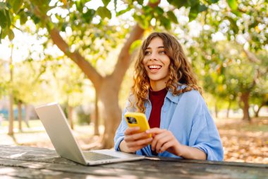 Genç gülümseyen öğrenci bahar parkında bankta oturur, dinlenir, dizüstü bilgisayar kullanır, cep telefonu interneti tarar. Kentsel yaşam tarzı kavramı.