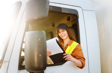 Genç kadın profesyonel kamyon şoförü bir otoparktaki sevkiyat listesini kontrol ediyor. Ulaştırma sektörü teması.