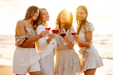 Kumsalda yürüyen şarap kadehli mutlu kadınlar. Bekarlığa veda partisini kutlamak, gün batımında şarap içmek. Tatiller, doğa, rahatlama konsepti.