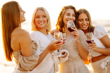 Kumsalda yürüyen şarap kadehli mutlu kadınlar. Bekarlığa veda partisini kutlamak, gün batımında şarap içmek. Tatiller, doğa, rahatlama konsepti.