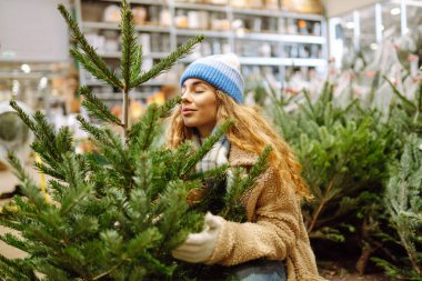 Sıcak giysiler içinde gülümseyen kadın markette bir Noel ağacı seçiyor. Kış tatili.