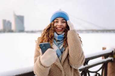 Mutlu bir kadın elinde telefonla karlı bir setin kenarında yürüyor. Çevrimiçi iletişim. Teknoloji konsepti.