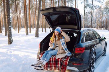 Mutlu Kadın bagajında oturuyor ve telefon kullanıyor. Karlı hava. Teknoloji kavramı, macera, arabayla seyahat..