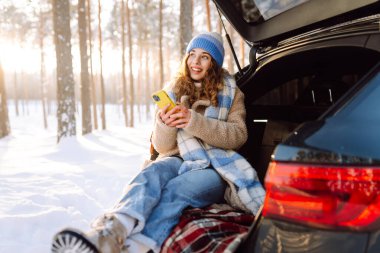Mutlu Kadın bagajında oturuyor ve telefon kullanıyor. Karlı hava. Teknoloji kavramı, macera, arabayla seyahat..
