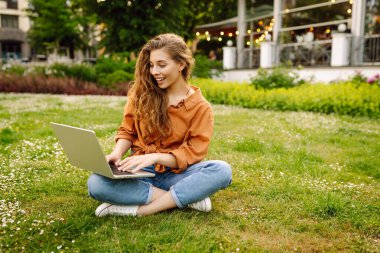 Genç kadın çimlerin üzerinde oturuyor, dizüstü bilgisayarla çalışıyor. Çevrimiçi eğitim, serbest çalışma, teknoloji konsepti. Mobil Ofis.