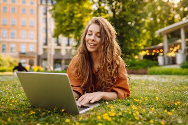 Genç kadın çimlerin üzerinde oturuyor, dizüstü bilgisayarla çalışıyor. Çevrimiçi eğitim, serbest çalışma, teknoloji konsepti. Mobil Ofis.
