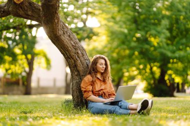 Genç kadın çimlerin üzerinde oturuyor, dizüstü bilgisayarla çalışıyor. Çevrimiçi eğitim, serbest çalışma, teknoloji konsepti. Mobil Ofis.