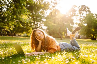 Genç kadın çimlerin üzerinde oturuyor, dizüstü bilgisayarla çalışıyor. Çevrimiçi eğitim, serbest çalışma, teknoloji konsepti. Mobil Ofis.