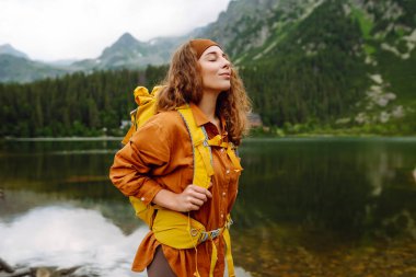 Sarı sırt çantalı genç turist kadın bir dağ gölünün arka planında duruyor. Seyahat, macera. Etkin bir yaşam biçimi kavramı.