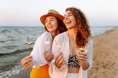 Güzel iki kadın sahilde maytap kutlaması yapıyor. Birlikte harika vakit geçiriyorlar! Yaz tatili, parti, tatil, dinlenme.