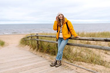 Sarı ceketli güzel bir kadın rüzgarlı bir sonbahar gününde sahilde dinleniyor. Doğa ve turizm kavramı.