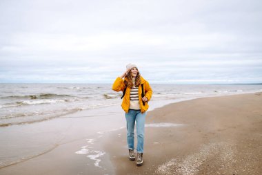 Sarı ceketli güzel bir kadın rüzgarlı bir sonbahar gününde sahilde dinleniyor. Doğa ve turizm kavramı.
